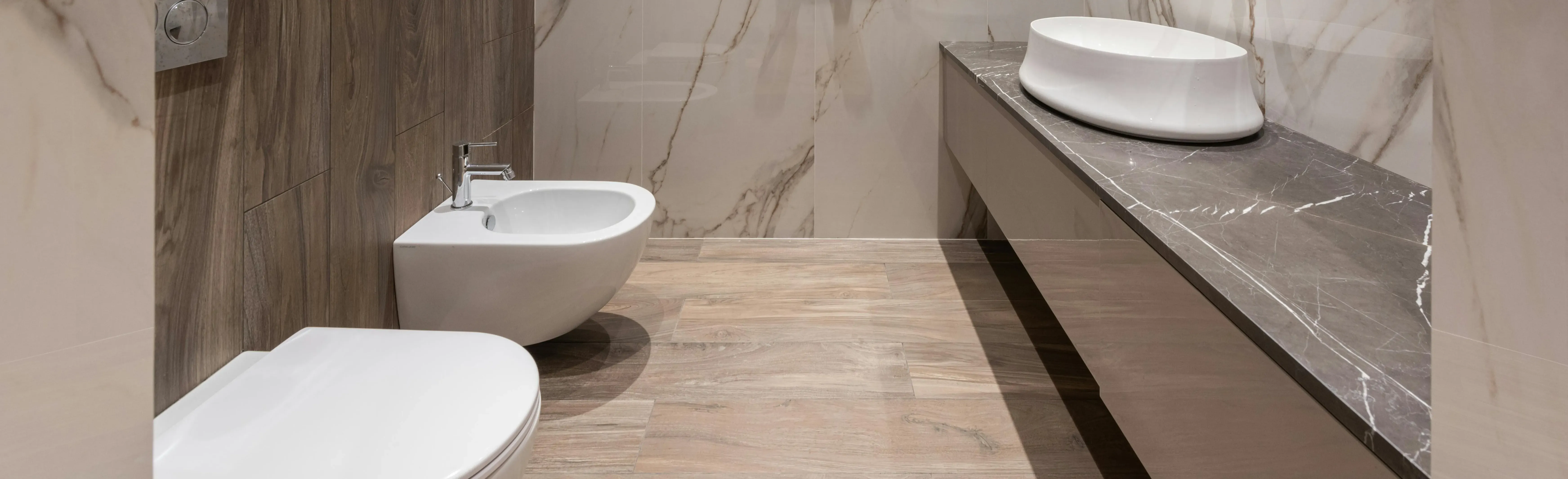 Detailaufnahme eines Wohnbads mit Vinylboden in Holzoptik, WC und Bidet vor einer Wand in heller Marmoroptik