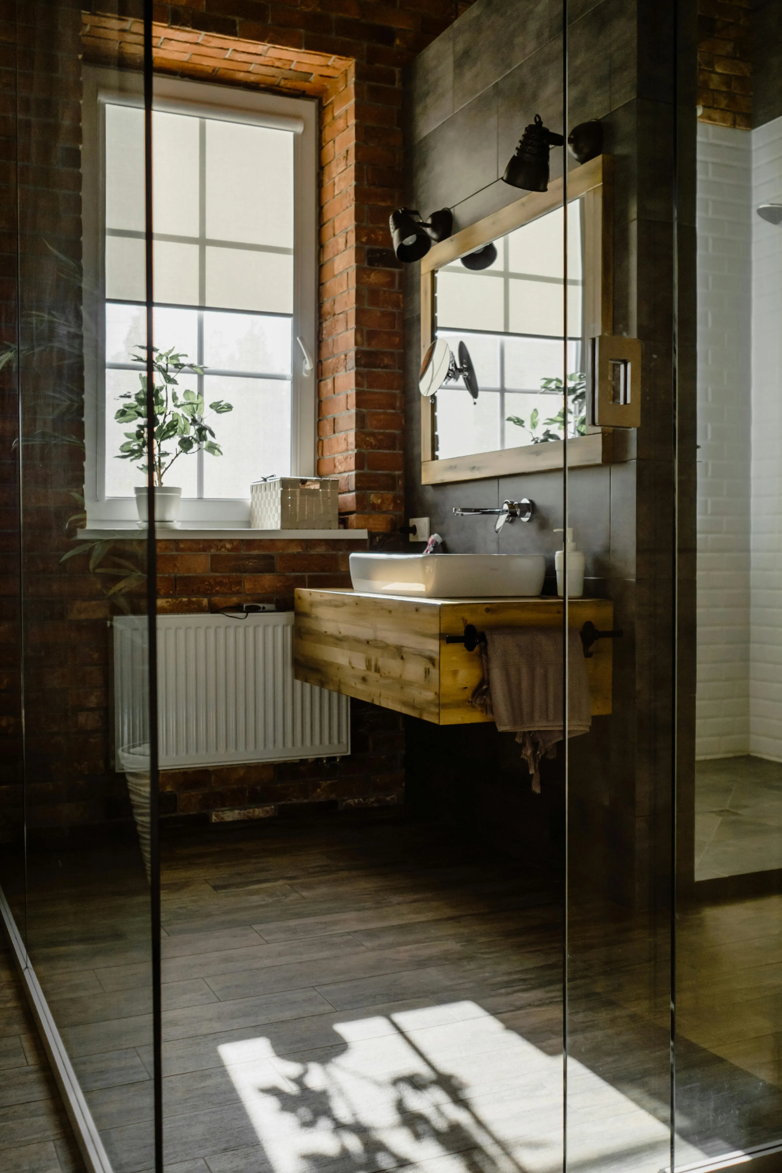 Modernes Industrial-Badezimmer mit Ziegelwand, dunklem Holzboden und bodengleicher Dusche hinter Glaswand als Grenzbereich für Vinylboden
