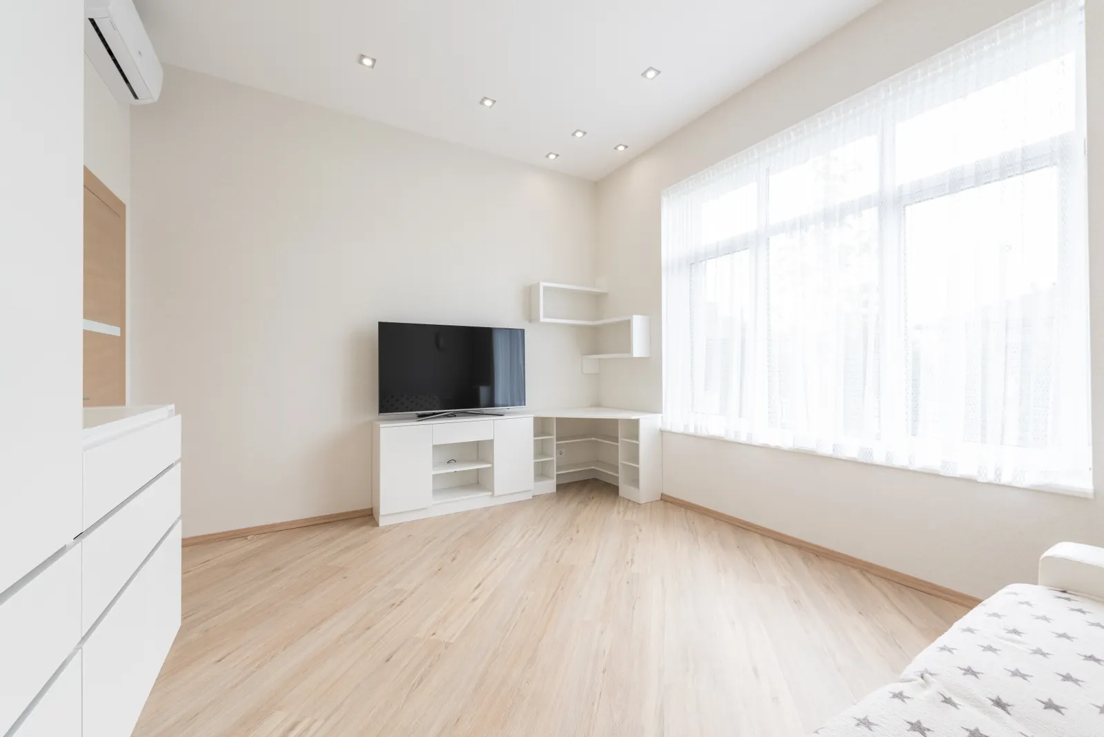 Heller Wohnraum mit hellem Holzoptik-Boden, weißem TV-Board, Kommode und großem Fenster mit Gardine
