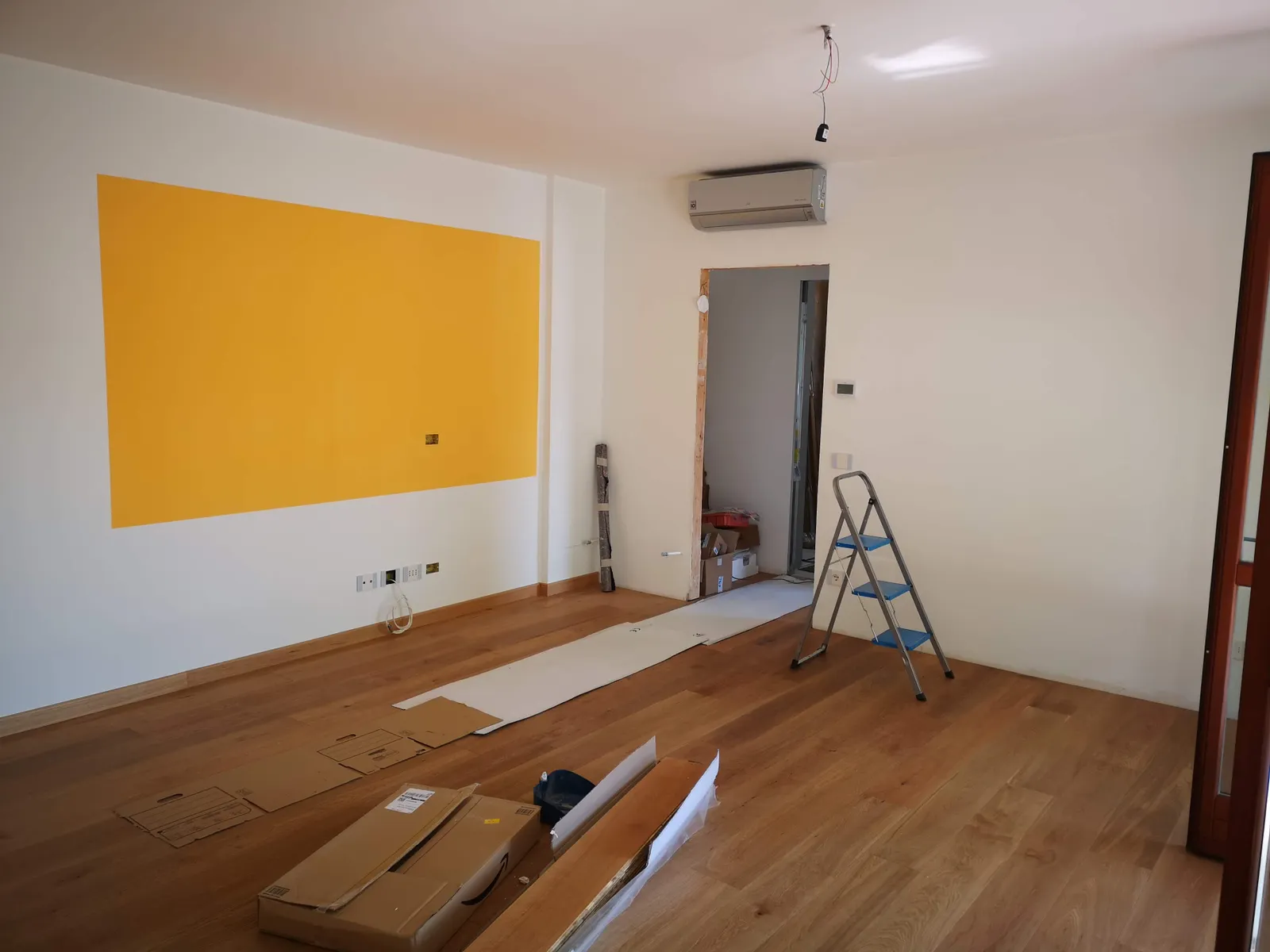 Raum in der Renovierung mit fertig verlegtem Dielenboden, teilweise gelb gestrichener Wand, Trittleiter und Kartons auf dem Boden